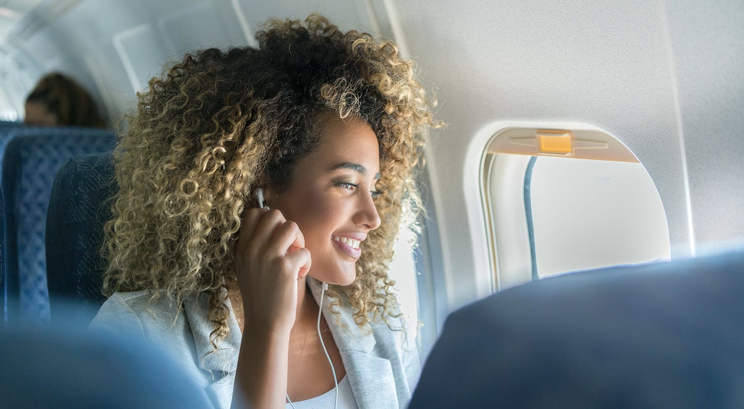 A woman with curly hair enjoys the view from her airplane window seat while wearing wired headphones.